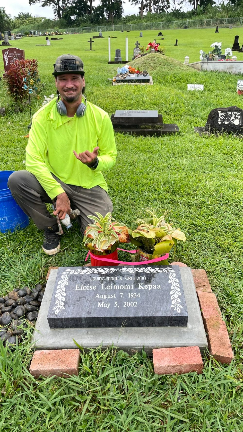 A man kneeling beside a flat memorial marker with flowers in a cemetery