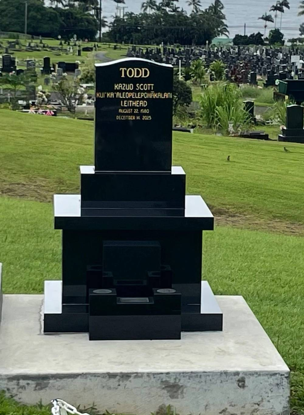 Black granite upright headstone with engraved name and gold lettering on a gray base, set on a green cemetery lawn in Hawaiʻi